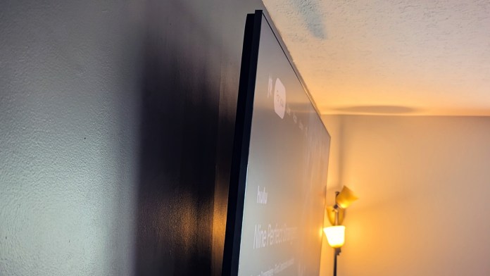 A close-up view of the side profile of a TCL 85QM6K TV, showcasing its thin design and modern finish next to a warm lamp and a wall.