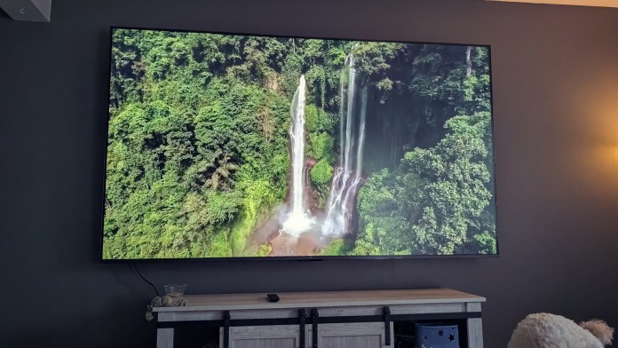 An 85-inch TCL TV displaying a scenic image of a waterfall surrounded by lush greenery, mounted on a wall in a modern living room setting.