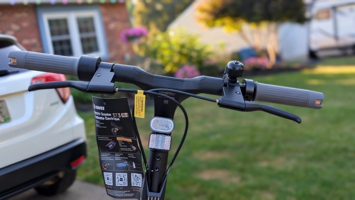 Close-up view of the handlebars of the Navee ST3 Pro electric scooter, featuring grips, brake lever, and a bell, with a yard and parked car in the background.