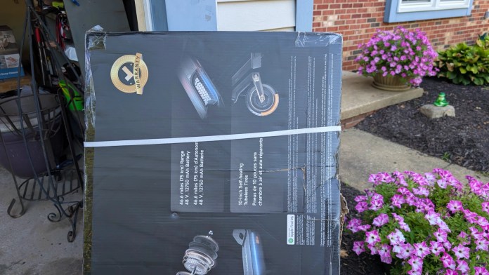 A partially opened box of the Navee ST3 Pro scooter featuring information on its specifications, including a warranty seal, range of 48.6 miles, and details about the battery and tire size, placed on a porch next to blooming flowers.