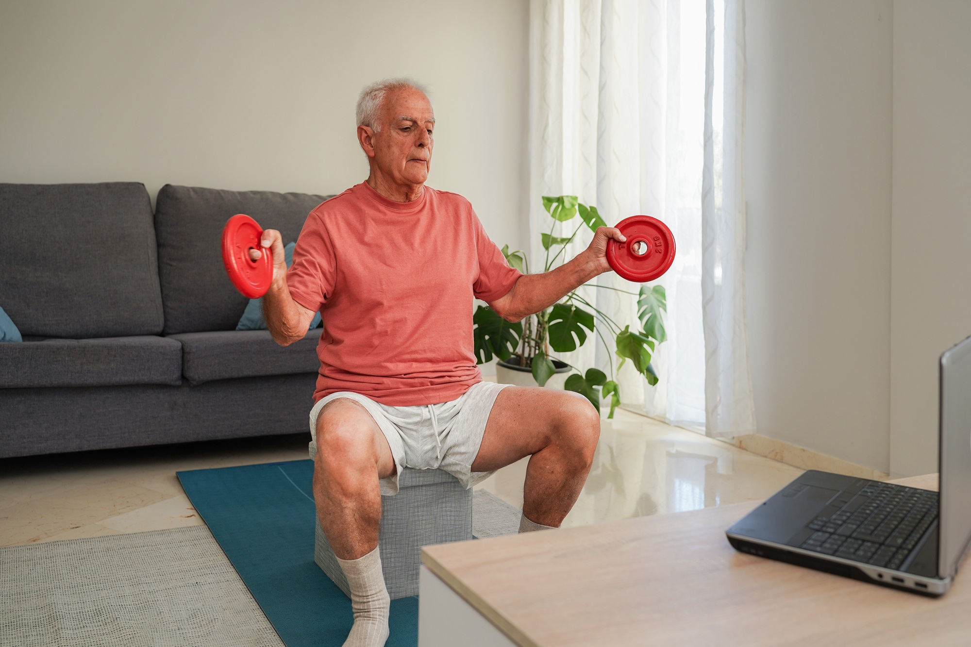 Senior man lifting weights at home following online training program on laptop