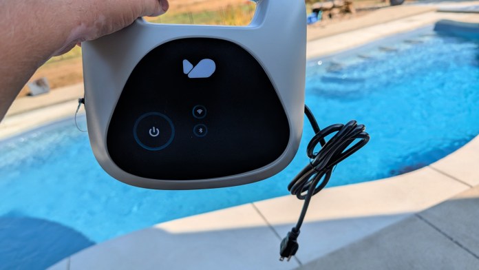 A hand holding the control panel of the Bublue BuHybrid L6 robotic pool cleaner, featuring buttons for power and connectivity, with a swimming pool in the background.