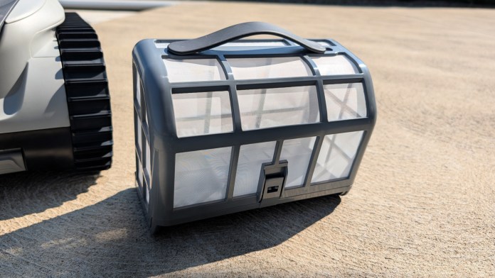 Close-up of a gray top-loading debris basket for a robotic pool cleaner, placed on a concrete surface next to the cleaner's wheel.
