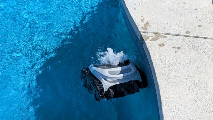 A robotic pool cleaner operating in a swimming pool, partially submerged in clear blue water, with water splashing around it.