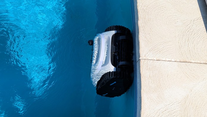 A robotic pool cleaner operating at the edge of a swimming pool, displaying its sleek design and black tracks in the clear blue water.