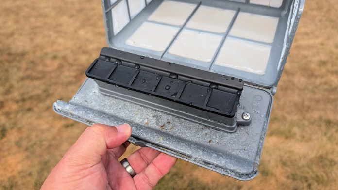 Close-up of a hand holding the filter basket of a robotic pool cleaner, showing its removable lid and water droplets, with a blurred grassy background.