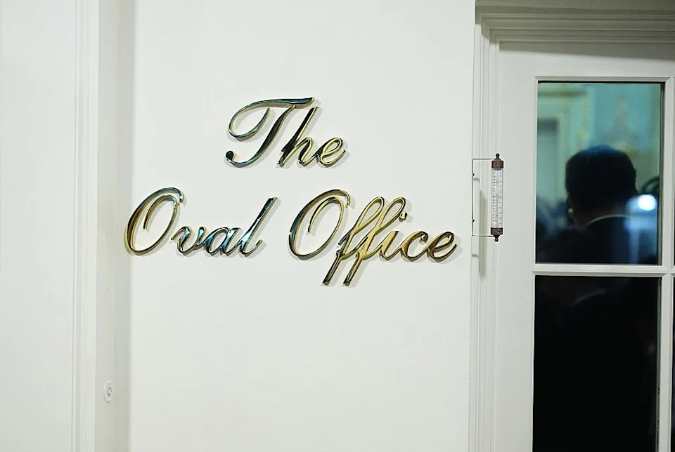 Sign reading "The Oval Office" on a wall, next to a window reflecting silhouettes of people