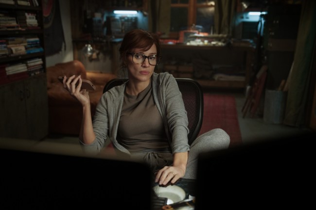 Jessica Chastain in 'The Savant,' shown sitting in an office chair, casually holding a cell phone, wearing gray sweats and black glasses