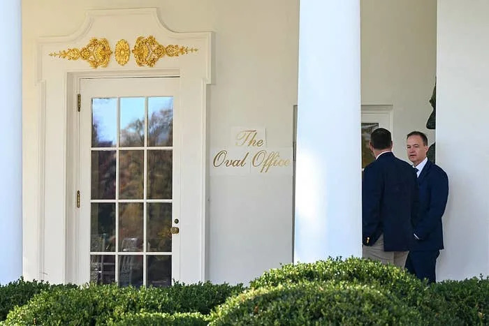 Two men in suits stand outside the Oval Office entrance, engaged in conversation